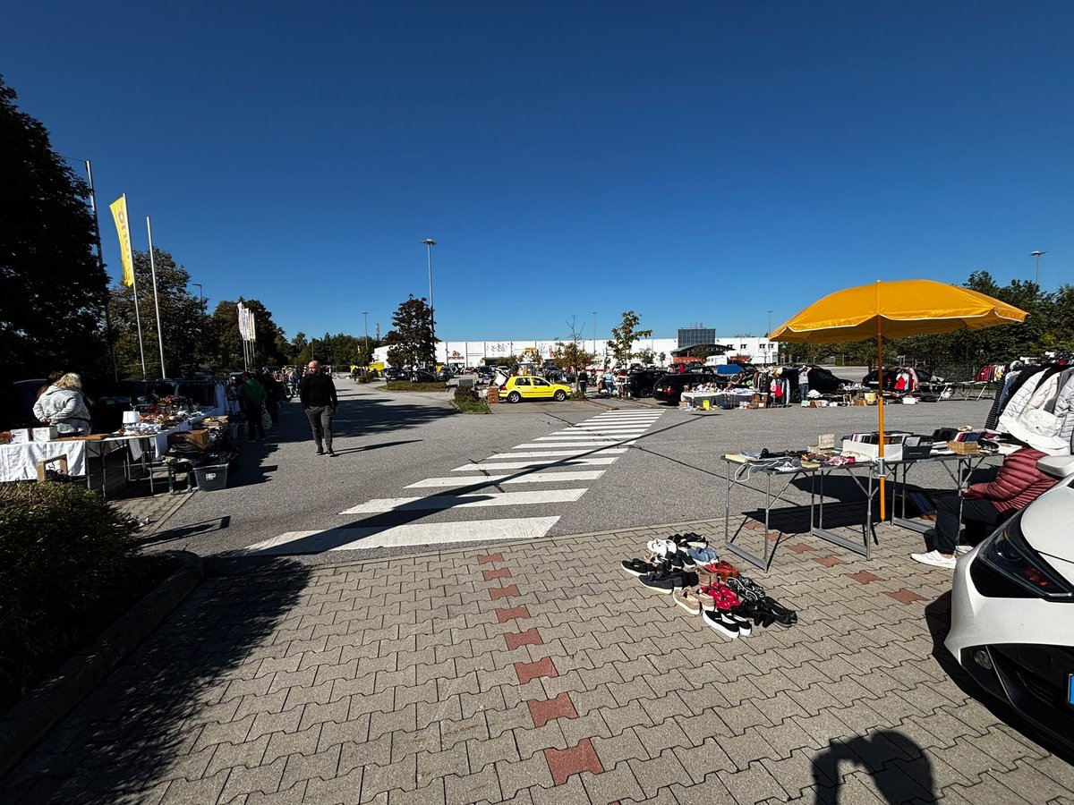 Flohmarkt mit Fahrzeugen, Verkaufszelten und gelbem Sonnenschirm auf dem Parkplatz.
